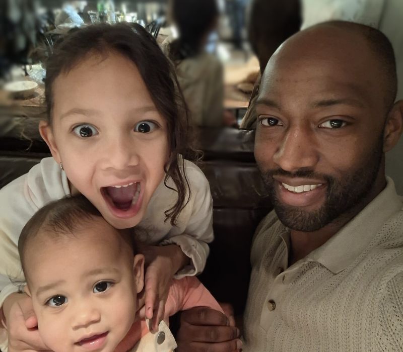 Een man glimlacht naar de camera terwijl hij een baby vasthoudt; naast hem leunt een jong meisje met grote ogen en open mond enthousiast naar hem toe. Ze zitten samen op een bank in het Amsterdamse restaurant Ysbreeker - vader Roms verbindt met zijn kinderen.