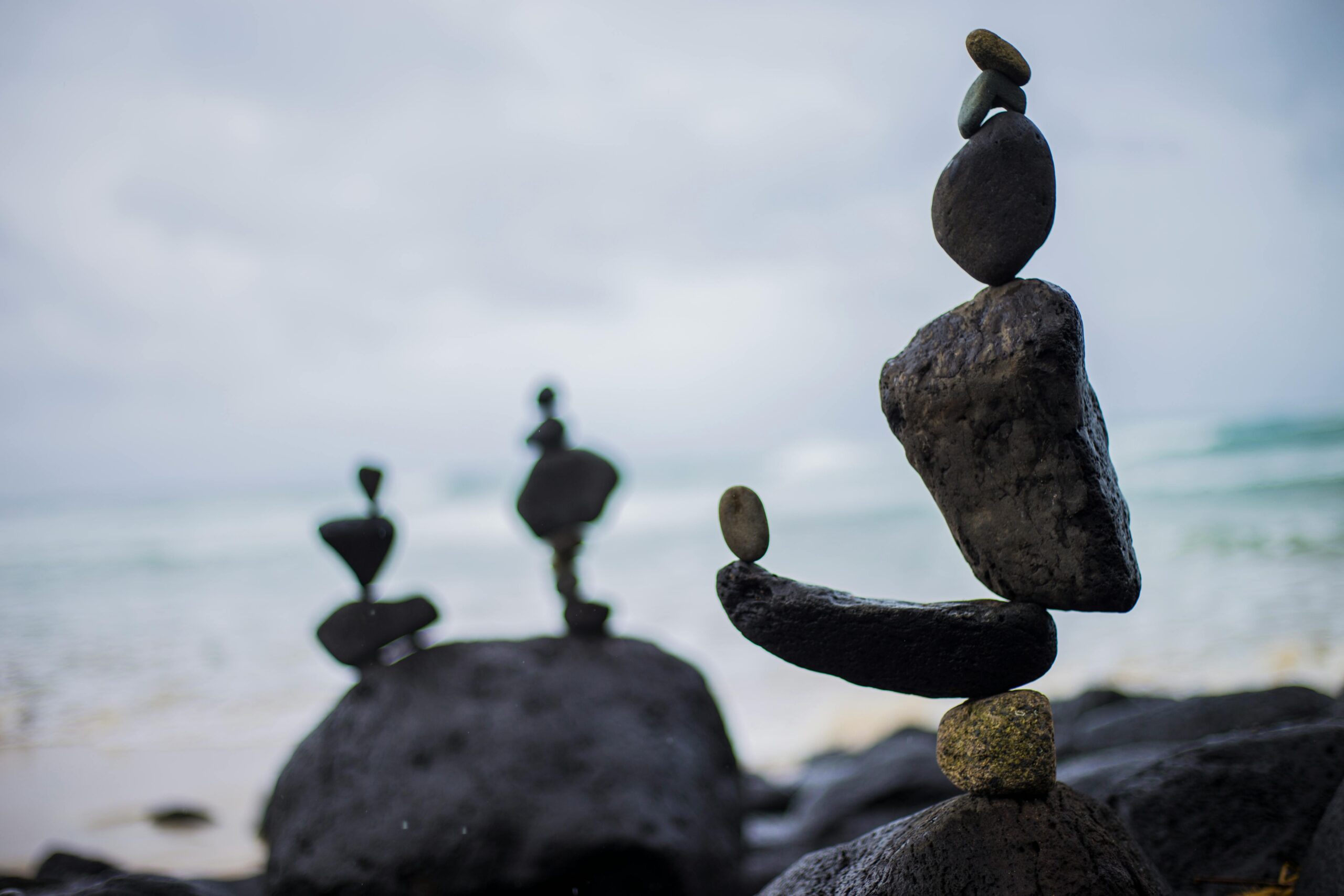 Close-up van zorgvuldig gestapelde stenen die een hoge toren vormen op het strand, met twee andere stenen stapels wazig op de achtergrond en de oceaangolven onder een bewolkte lucht.